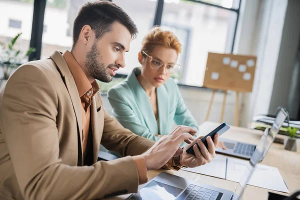 Bärtiger Geschäftsmann mit Smartphone in der Nähe von Laptops und Frau mit Brille im Büro — Stockfoto