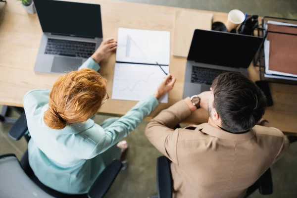 Вид сверху рыжеволосой бизнесвумен, указывающей на аналитику во время работы рядом с ноутбуками и коллегой в офисе — стоковое фото