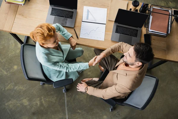 Вид зверху бізнес-партнерів, що тремтять руками біля ноутбуків і документів з графіками в офісі — стокове фото