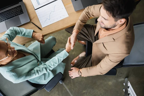 Top view of happy partners shaking hands near laptops and business analytics in office — Stock Photo