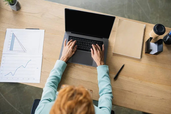 Вид сверху на рыжую деловую женщину, работающую на ноутбуке рядом с бумагами с графиками в офисе — стоковое фото