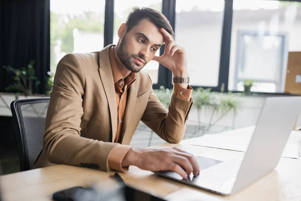 Hombre de negocios aburrido en chaqueta beige mirando a la computadora portátil mientras está sentado en el lugar de trabajo en la oficina - foto de stock