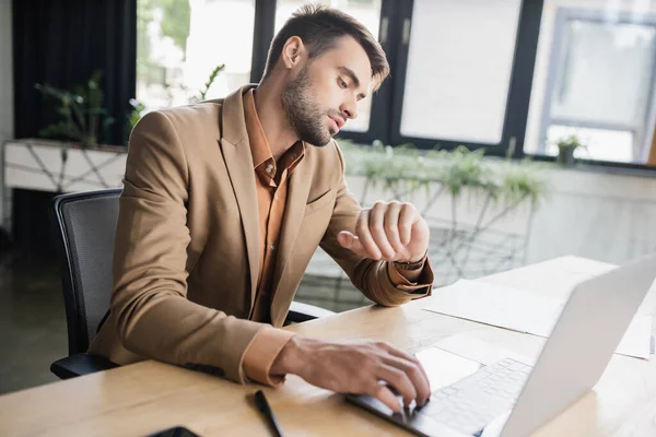 Homem de negócios barbudo em blazer elegante verificando o tempo enquanto sentado no laptop no escritório — Fotografia de Stock