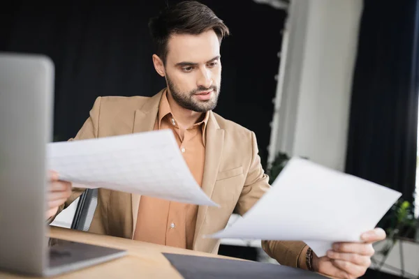 Bearded businessman looking at papers while working near blurred laptop in office — Stock Photo