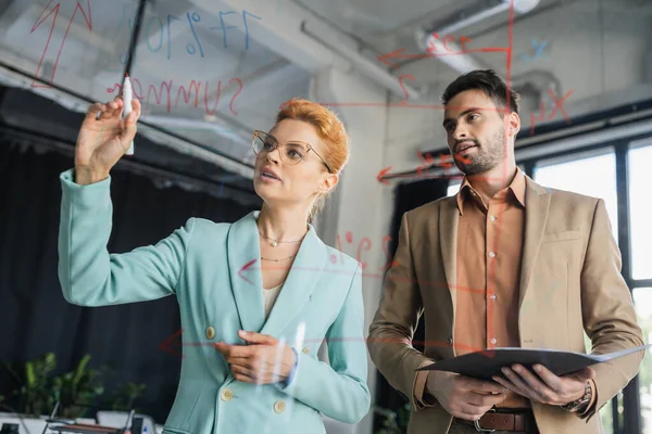 Рыжая деловая женщина в очках указывает на знаки на стеклянной доске рядом с коллегой с папкой в офисе — стоковое фото
