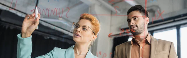 Businesswoman with felt pen writing on glass board near bearded colleague in office, banner — Stock Photo