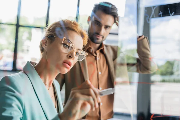 Thoughtful woman in eyeglasses writing on glass board near young businessman in office — Stock Photo