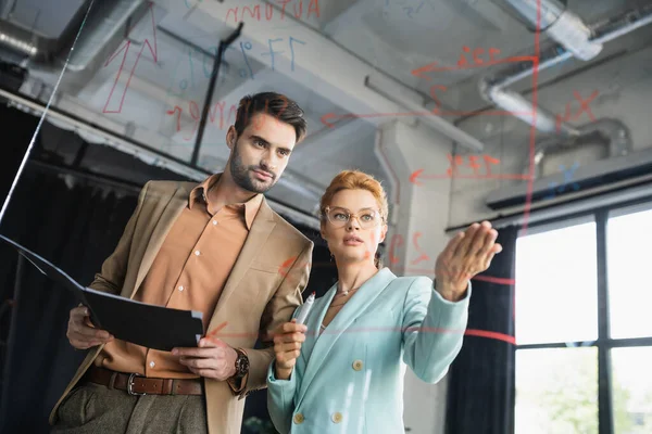 Redhead businesswoman pointing at lettering on glass board near manager with folder in office — Stock Photo