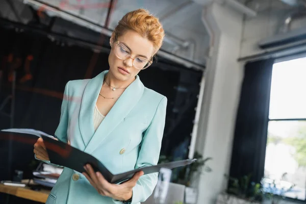 Pensativo pelirroja mujer de negocios en gafas y elegante chaqueta mirando carpeta con documentos en la oficina - foto de stock