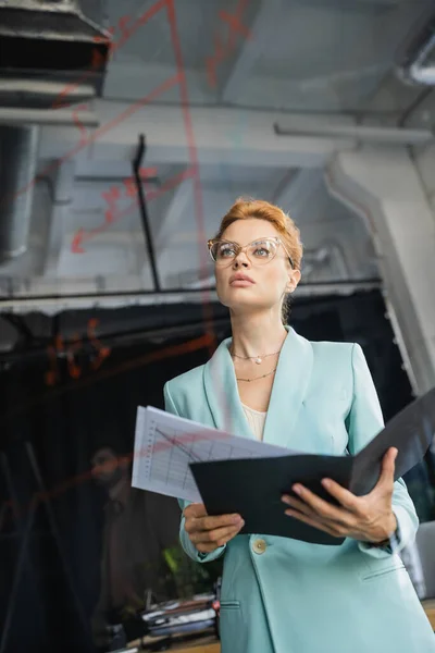 Tiefansicht einer rothaarigen Geschäftsfrau mit Brille, die Dokumente hält und im Büro Schilder auf Glasplatten betrachtet — Stockfoto