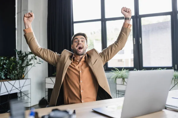 Overjoyed businessman looking at camera while shouting and rejoicing near laptop in office — Stock Photo