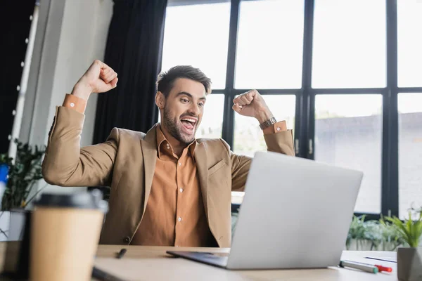 Joyful businessman looking at laptop and showing triumph gesture in office — Stock Photo