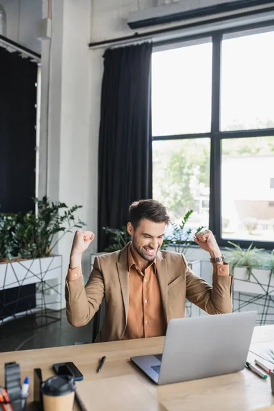 Excited businessman showing triumph gesture while sitting near laptop in office — Stock Photo