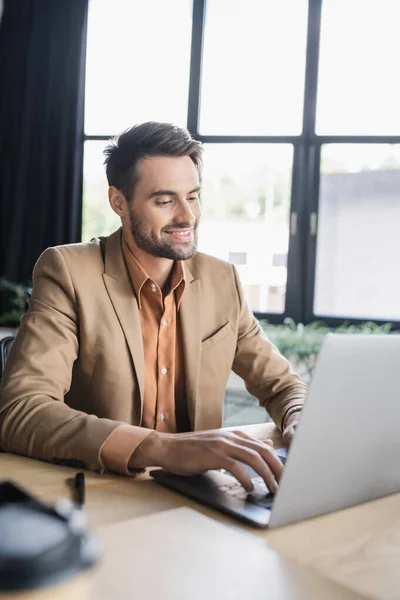 Улыбающийся бизнесмен в стильном пиджаке, печатающий на ноутбуке во время работы в современном офисе — стоковое фото