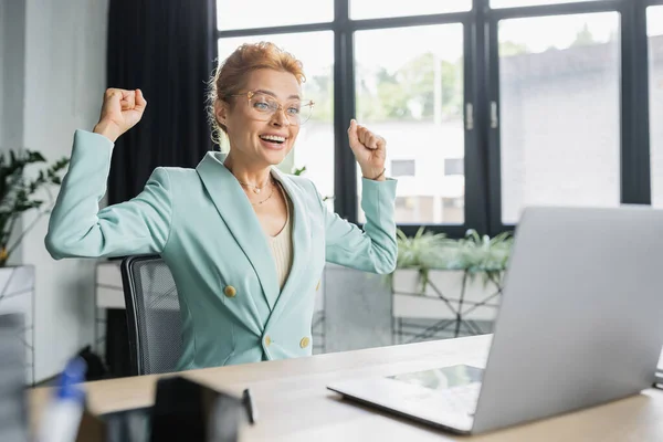 Cheerful redhead businesswoman in eyeglasses showing win gesture near laptop in office — Stock Photo