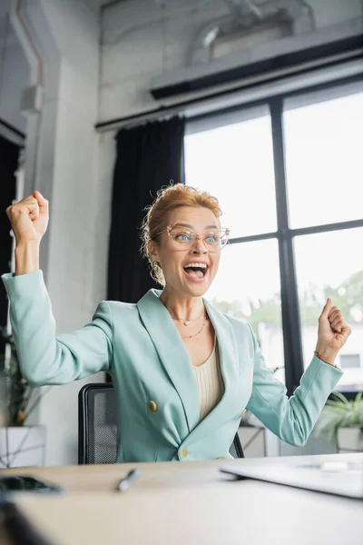 Overjoyed redhead businesswoman in eyeglasses showing win gesture and screaming in office — Stock Photo