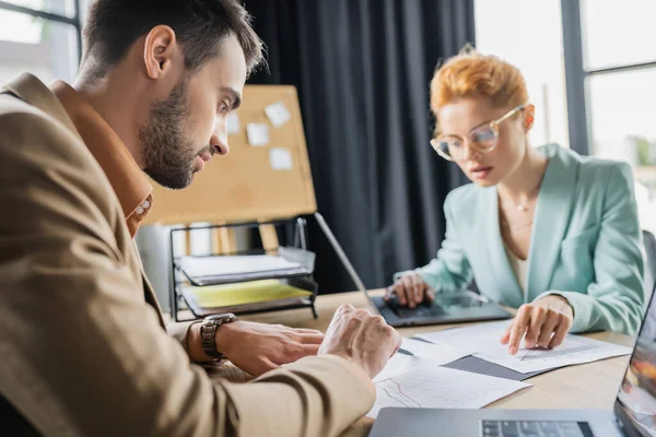 Blurred businesswoman in eyeglasses pointing at documents while working near laptop and bearded colleague in office — Stock Photo