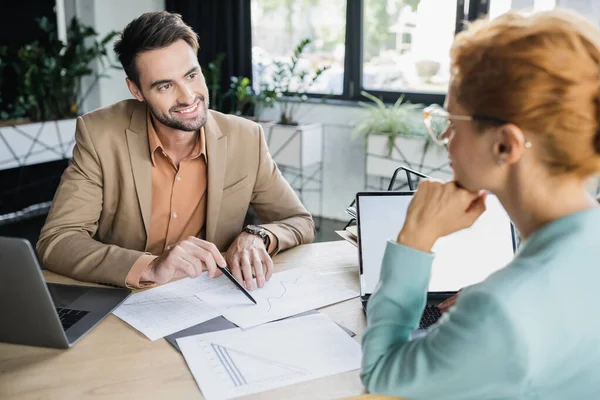 Smiling bearded manager pointing at charts near businesswoman on blurred foreground in office — Stock Photo