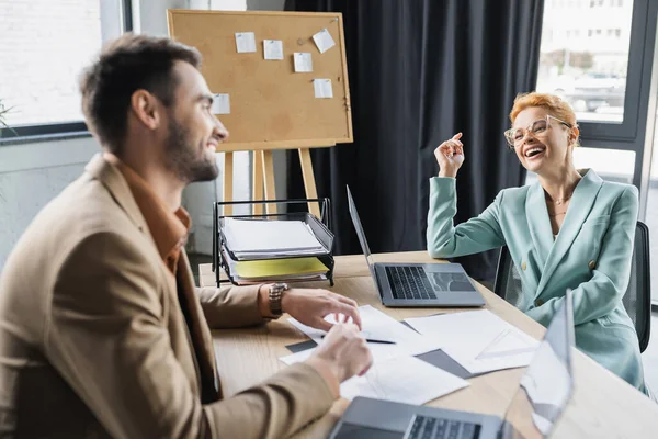 Збуджена бізнес-леді в окулярах сміється біля розмитого колеги на робочому місці в офісі — стокове фото