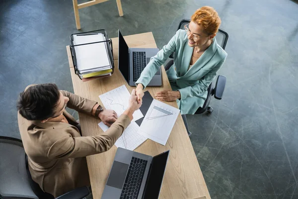 Top view of successful partners shaking hands near business analytics and laptops in office — Stock Photo