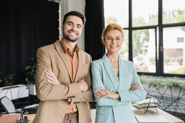 Erfolgreiche Geschäftskollegen in stylischen Anzügen stehen mit verschränkten Armen da und lächeln in die Kamera im Büro — Stockfoto
