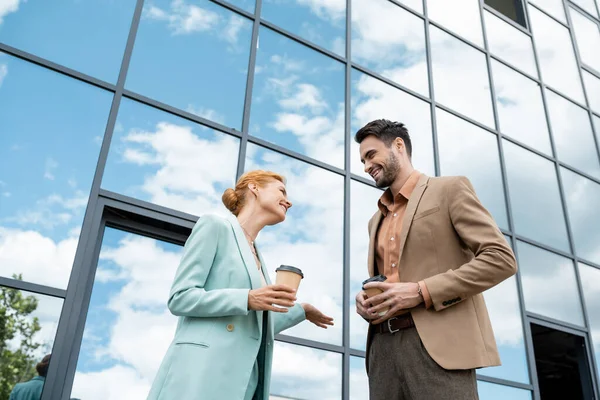 Низкий угол обзора довольных деловых партнеров с кофе идти говорить рядом с современным зданием со стеклянным фасадом — стоковое фото