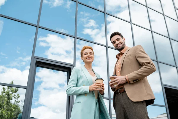 Low angle view of cheerful and trendy managers with coffee to go smiling at camera near glass building on city street — Stock Photo