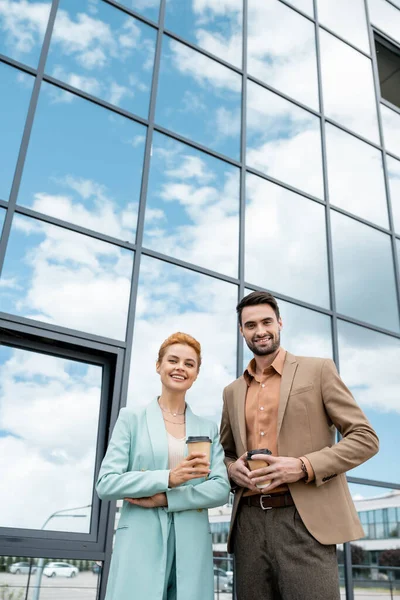 Hommes d'affaires élégants avec des tasses en papier souriant à la caméra près du bâtiment moderne avec façade en verre — Photo de stock