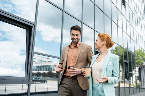 Compañeros de negocios alegres con bebidas para llevar hablando mientras camina por el edificio moderno en la calle urbana - foto de stock