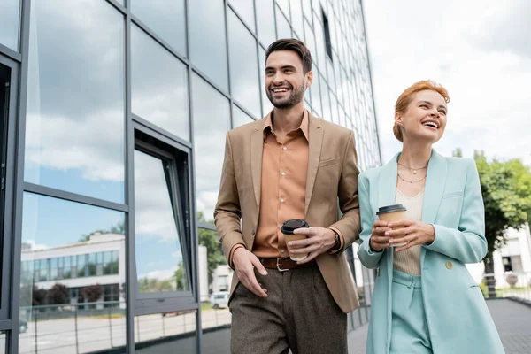 Cheerful business people in trendy clothes walking with coffee to go on urban street — Stock Photo