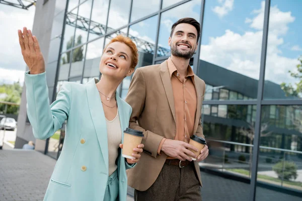 Happy redhead businesswoman looking away and pointing with hand while walking with colleague on city street — Stock Photo