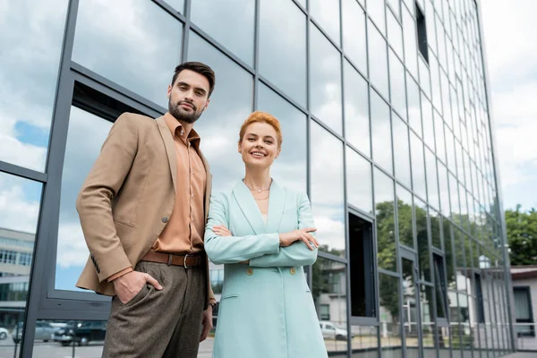 Happy and stylish managers looking at camera while posing near modern building on city street — Stock Photo