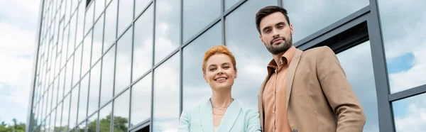 Позитивные деловые партнеры в стильных пиджаках, смотрящие в камеру возле здания города со стеклянным фасадом, баннером — стоковое фото