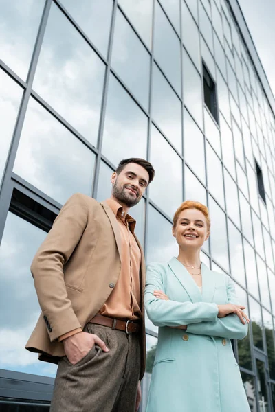 Low angle view joyful and trendy business partners looking at camera while posing near glass building in city — Stock Photo