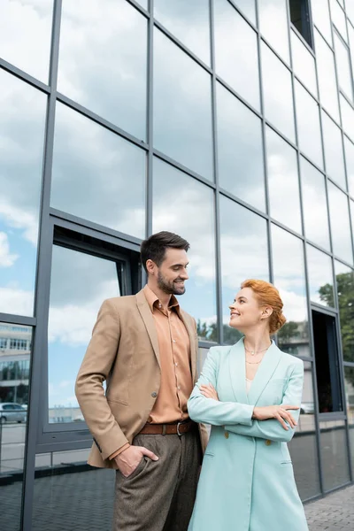 Stylish business people in blazers smiling at each other near urban building with glass facade — Stock Photo
