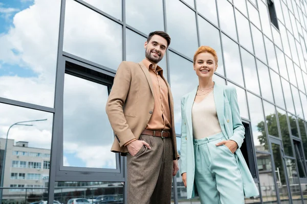 Gens d'affaires souriants dans des vêtements élégants posant avec les mains dans les poches près du bâtiment en verre sur la rue urbaine — Photo de stock