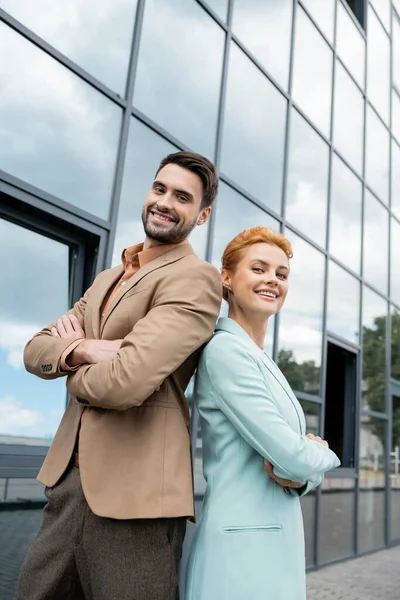 Collègues d'affaires joyeux posant dos à dos près du bâtiment en verre et souriant à la caméra — Photo de stock