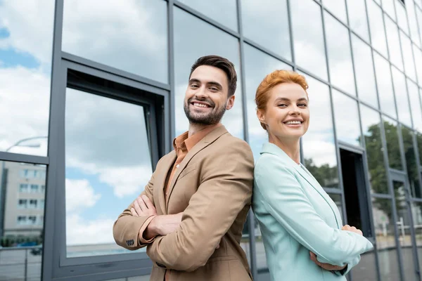 Happy business partners posing back to back with folded arms near modern building on urban street — Stock Photo