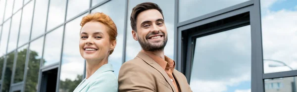 Счастливые и стильные деловые партнеры, улыбающиеся перед камерой возле стеклянного здания на городской улице, баннер — стоковое фото