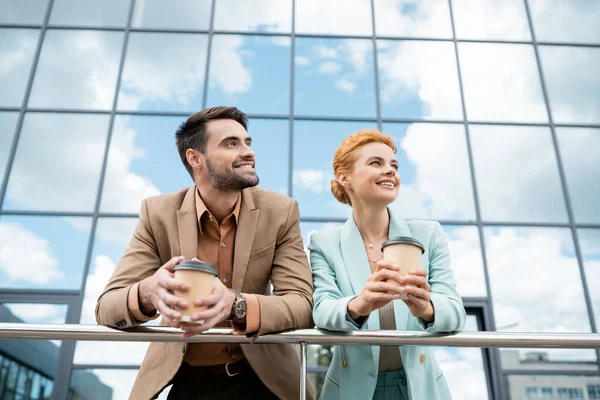 Joyful managers in stylish blazers holding paper cups and looking away near railing and modern building on urban street — Stock Photo