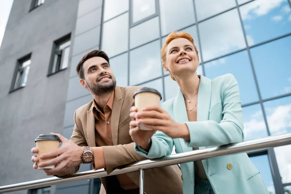 Lächelnde Geschäftspartner in trendigen Klamotten halten Coffee to go in der Hand und schauen am Geländer der städtischen Straße weg — Stockfoto