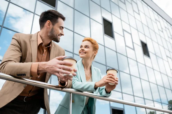 Pleased business partners in trendy clothes holding coffee to go and looking at each other near railing on urban street — Stock Photo