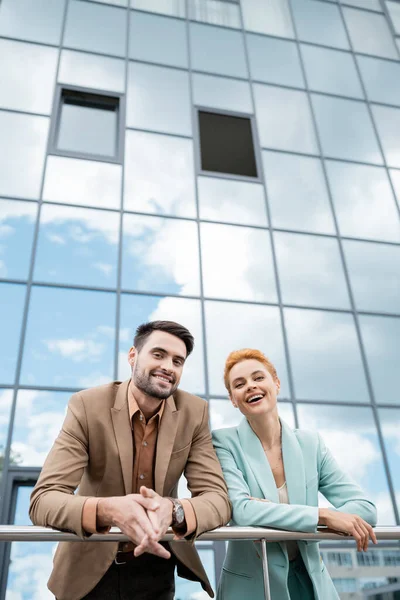 Cheerful and trendy managers smiling at camera near railing and city building with glass facade — Stock Photo
