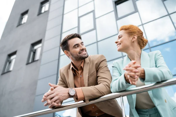Low angle view smiling business partners in stylish jackets looking at each other near railing on urban street — Stock Photo