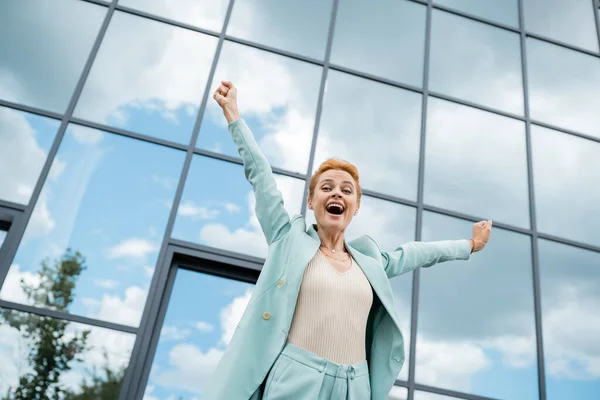 Vista de ángulo bajo de la mujer de negocios emocionada mostrando gesto de triunfo y gritando cerca del edificio de vidrio en la ciudad - foto de stock