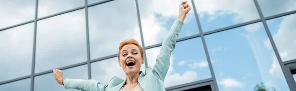 Low angle view of cheerful redhead businesswoman celebrating triumph near modern building in city, banner — Stock Photo