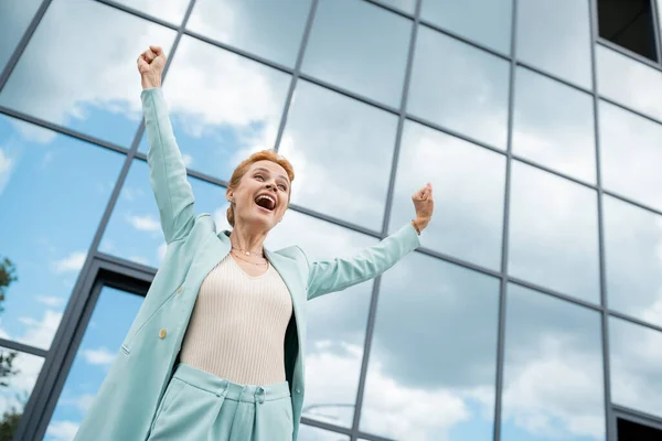 Vista de ángulo bajo de la mujer de negocios llena de alegría que muestra el gesto de ganar y gritando cerca del edificio con fachada de vidrio - foto de stock