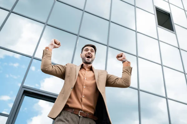 Vista de ángulo bajo de empresario alegre gritando y mostrando gesto de éxito cerca de edificio urbano con fachada de vidrio - foto de stock