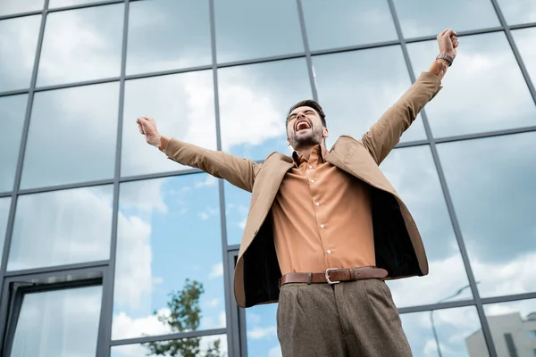 Low angle view of successful overjoyed businessman in trendy clothes showing triumph gesture near modern city building — Stock Photo