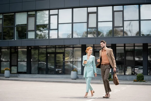 Full length of trendy business partners with paper cups walking near modern building on urban street — Stock Photo